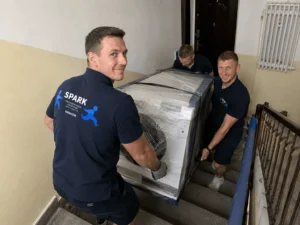 Pracownicy firmy przeprowadzkowej przenoszą zabezpieczoną pralkę w mieszkaniu