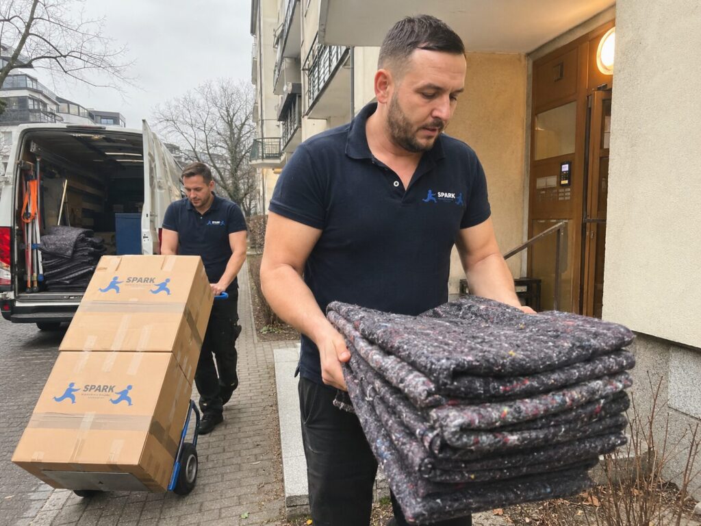 Pracownik firmy przeprowadzkowej przenosi zabezpieczony mebel do samochodu dostawczego.