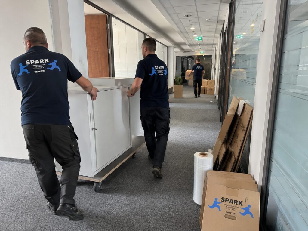 Pracownicy Spark Przeprowadzki przenoszą zabezpieczony mebel przez korytarz budynku.